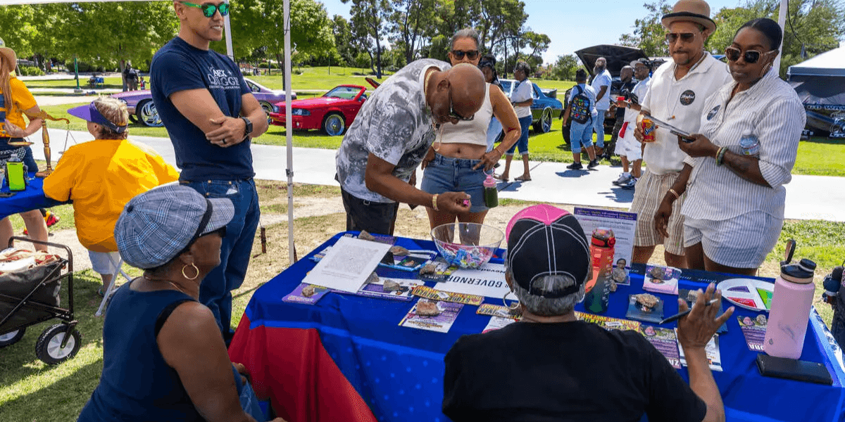 People interacting at an outdoor event with a colorful vendor booth and cars in the background