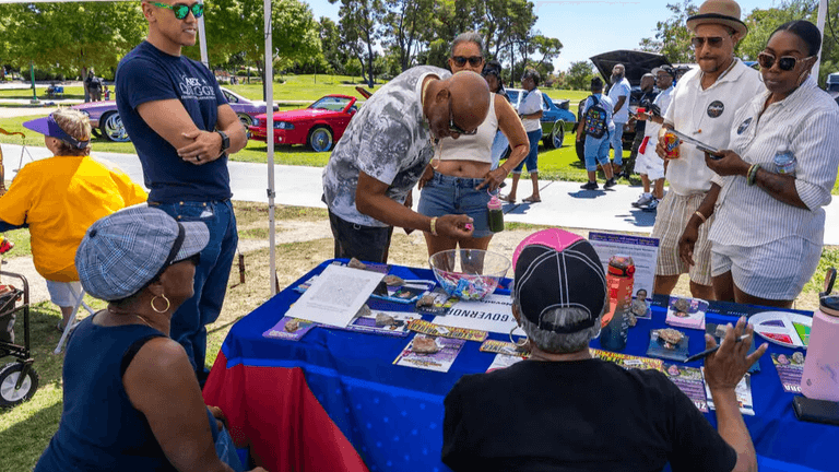 People interacting at an outdoor event with a colorful vendor booth and cars in the background