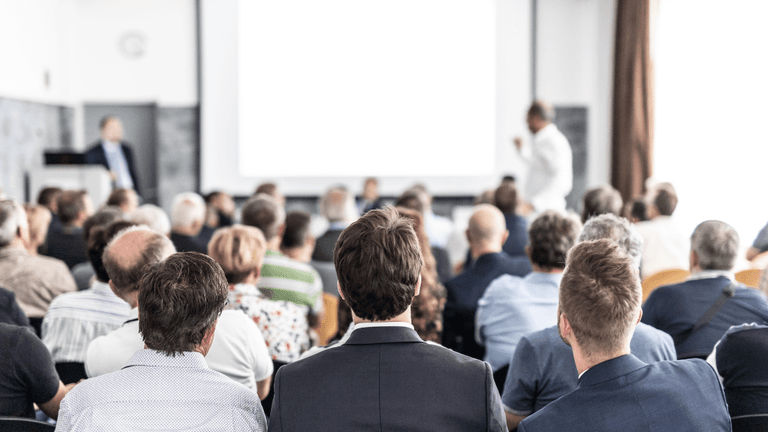 Audience facing a speaker at a presentation in a well-lit room