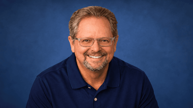 Smiling man with glasses wearing a navy polo shirt against a blue background