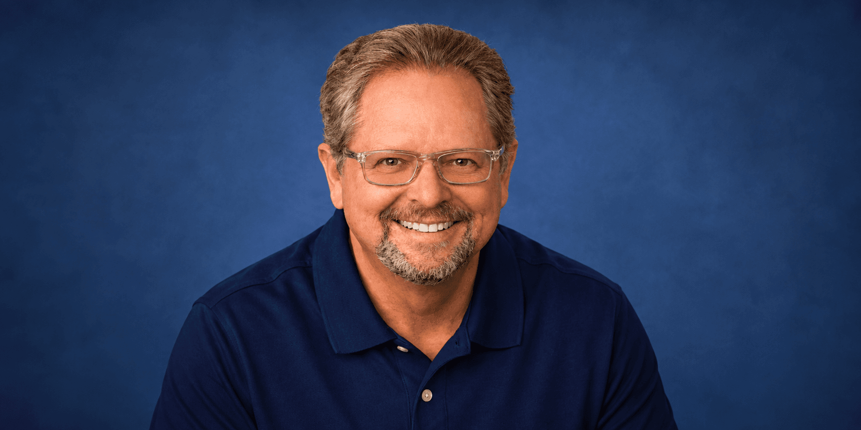 Smiling man with glasses wearing a navy polo shirt against a blue background