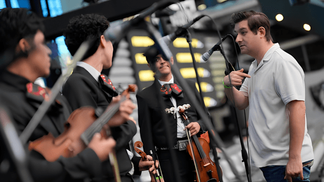 A man speaks to a mariachi band performing indoors with instruments in hand
