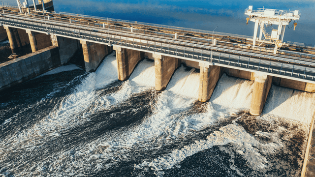 Water flows through open gates of a large dam structure with industrial equipment spanning the reservoir.