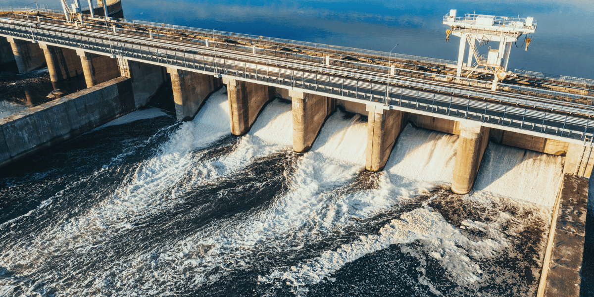 Water flows through open gates of a large dam structure with industrial equipment spanning the reservoir.